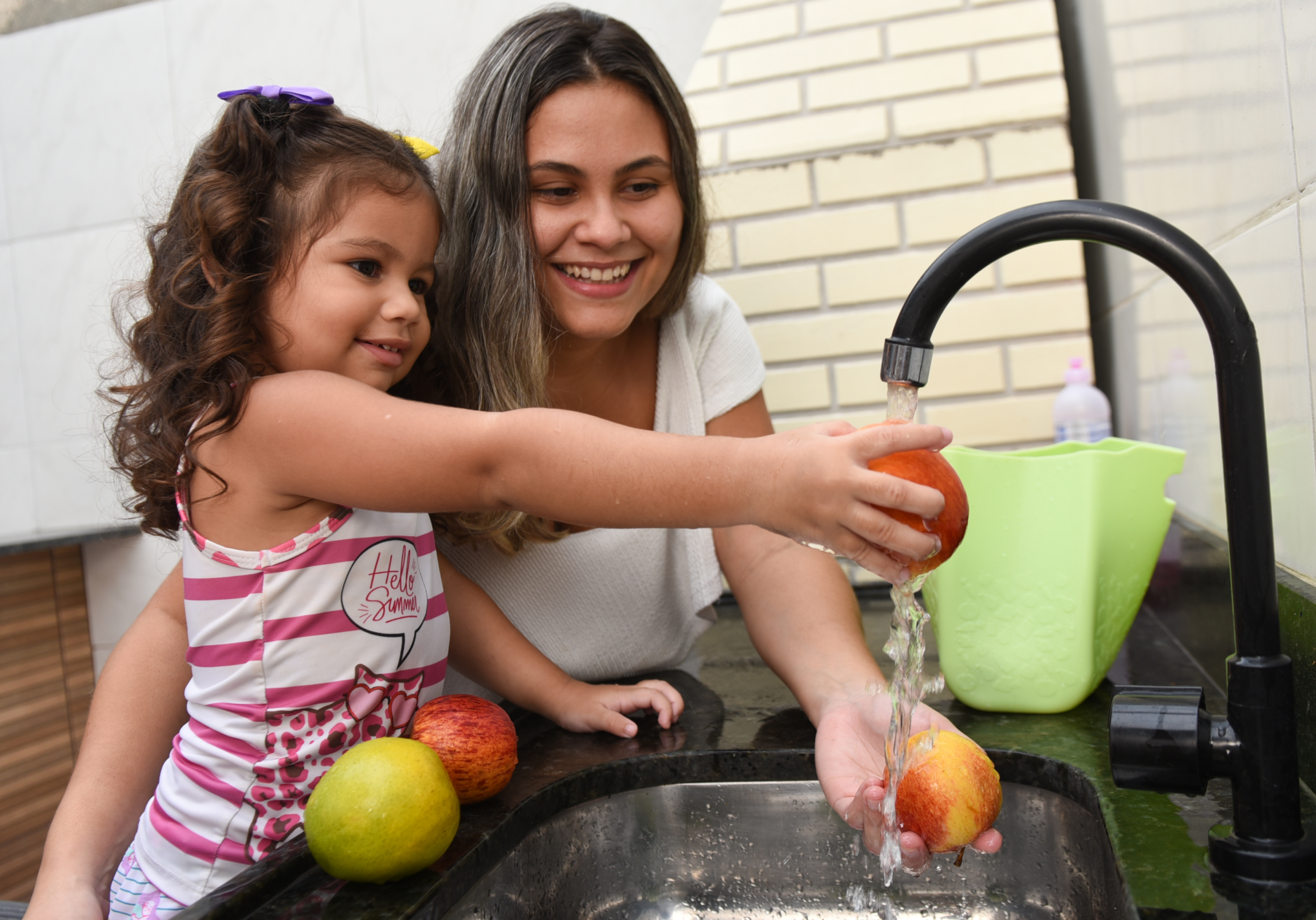 Mãe e filha lavam maçãs e laranjas na torneira de casa.
